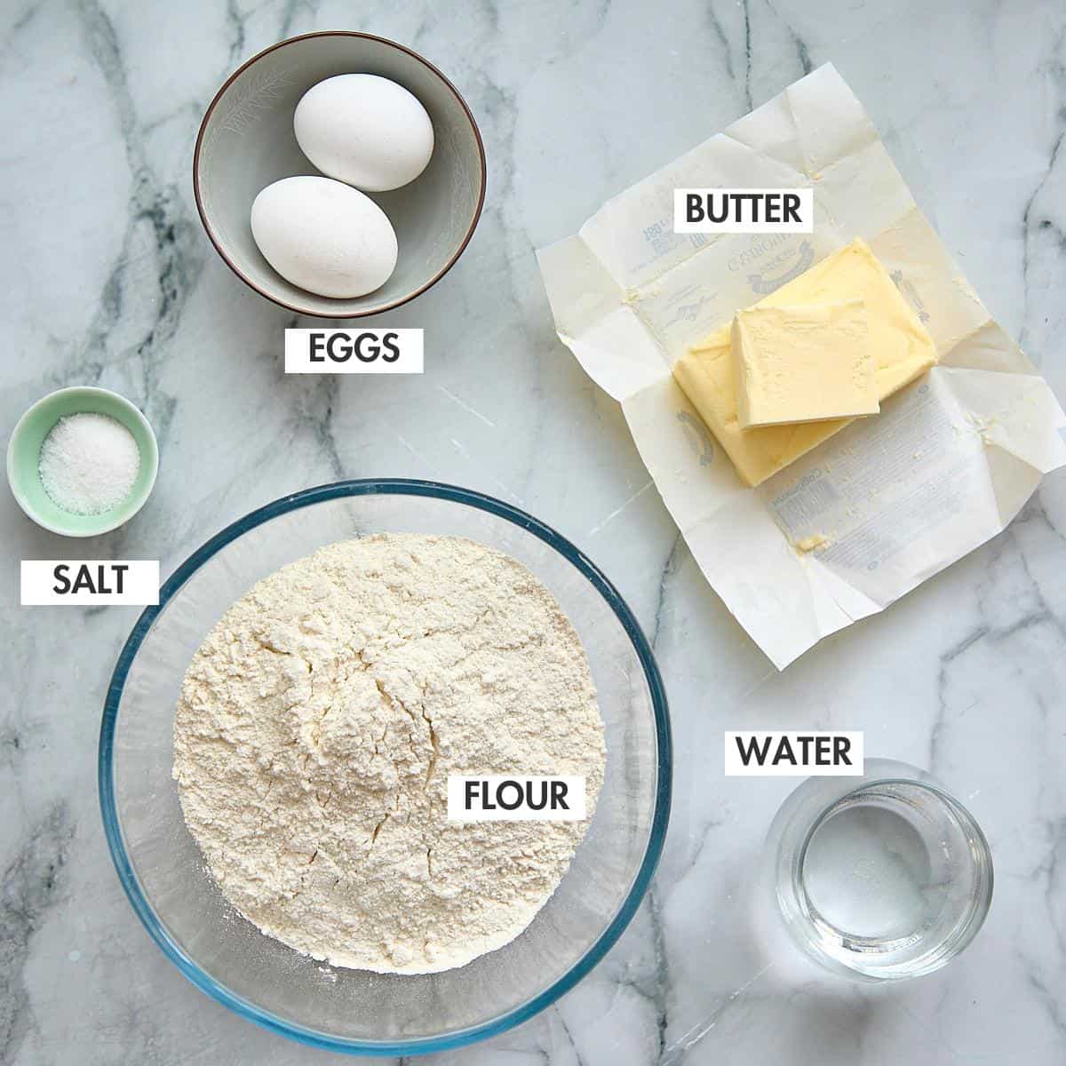 Two eggs in a ceramic bowl, butter sticks unwrapped, flour in a glass bowl, salt in a small dish, and water in a glass cup arranged on marble.