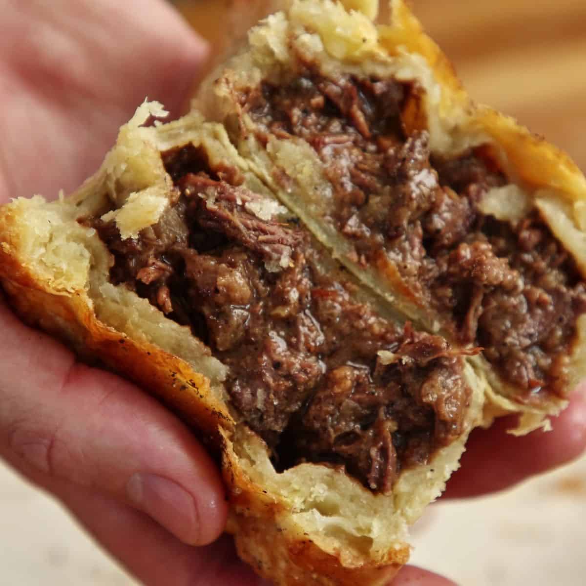 Single pepper steak pie cut in half on white plate showing glossy pepper-flecked beef filling with visible chunks and golden flaky pastry layers.