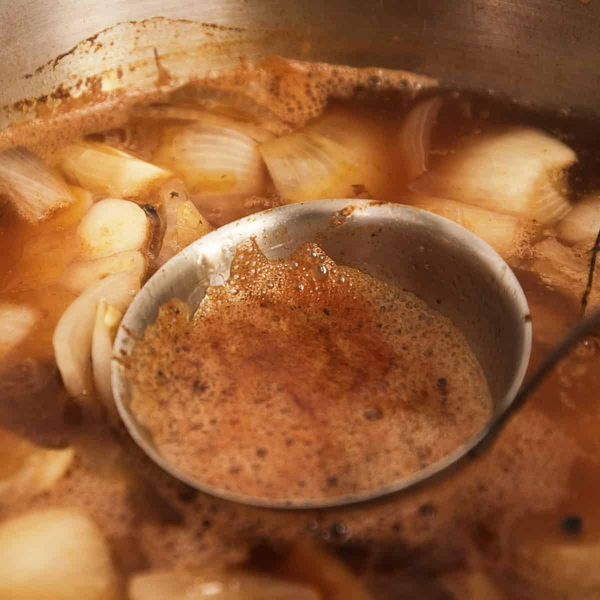 Using a ladle to skim fat and froth off beef stock while cooking in a stock pot.