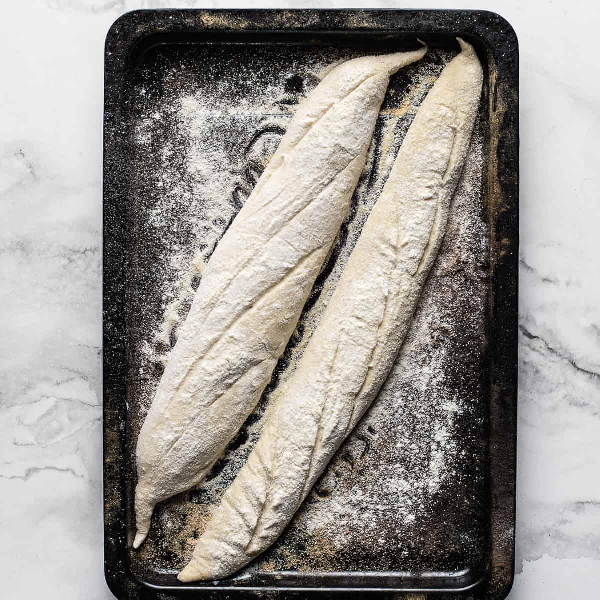 two raw baguettes slashed on a black oven tray