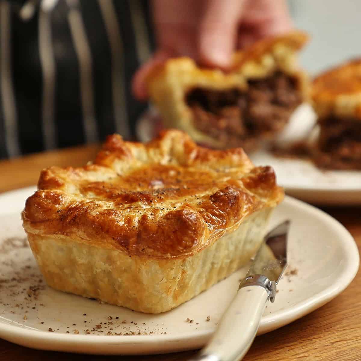 South African pepper steak pie on a white plate with a cross section of a pie cut in half in the background.