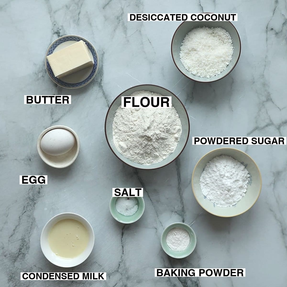 Tennis biscuit ingredients arranged on white surface: cake flour, desiccated coconut, condensed milk, butter, egg, baking powder