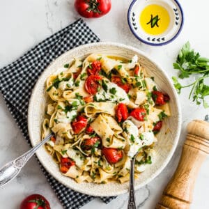 cherry tomato pasta in white bowl served on a black napkin with tomato, olive oil, pepper grinder and parsley in the background