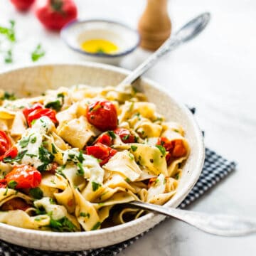 cherry tomato pasta in white bowl served on a black napkin with tomato, olive oil, pepper grinder and parsley in the background
