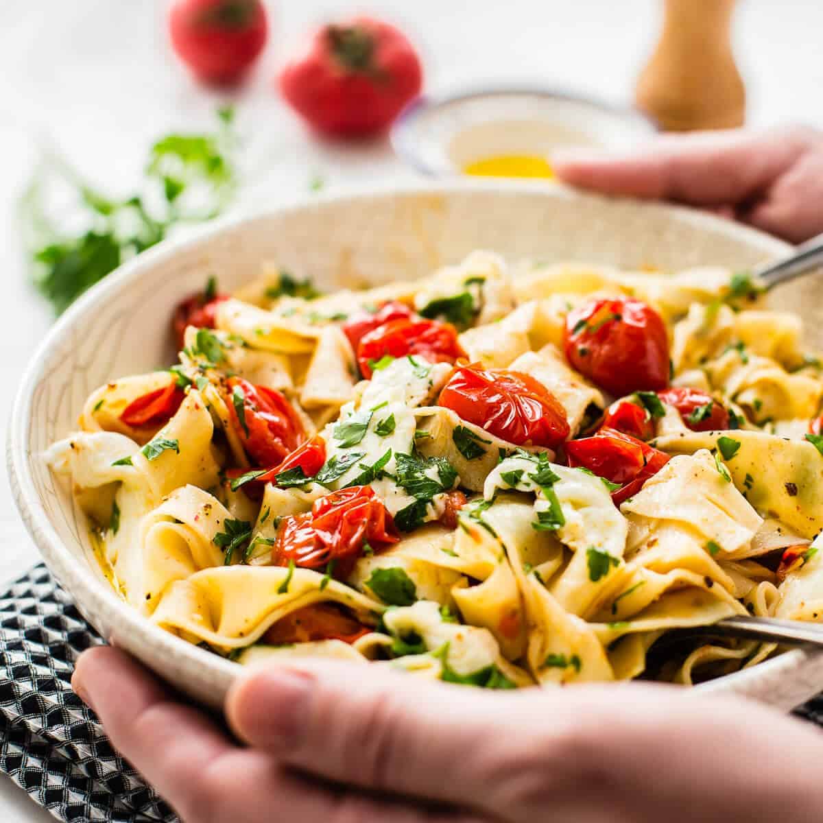 cherry tomato pasta in white bowl. Hands holding the bowl served on a black napkin with tomato, olive oil, pepper grinder and parsley in the background