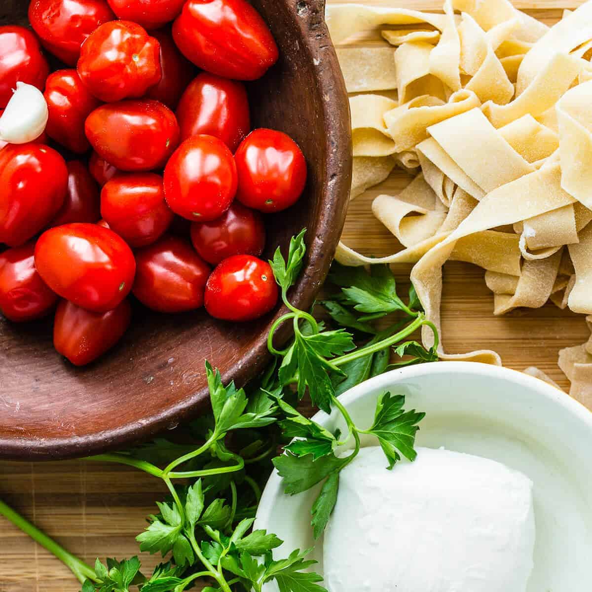 red cherry tomatoes in brown bowl on wooden chopping board with freshly made raw tagliatelle pasta, green parsley and fresh buffalo mozzarella ball in white bowl