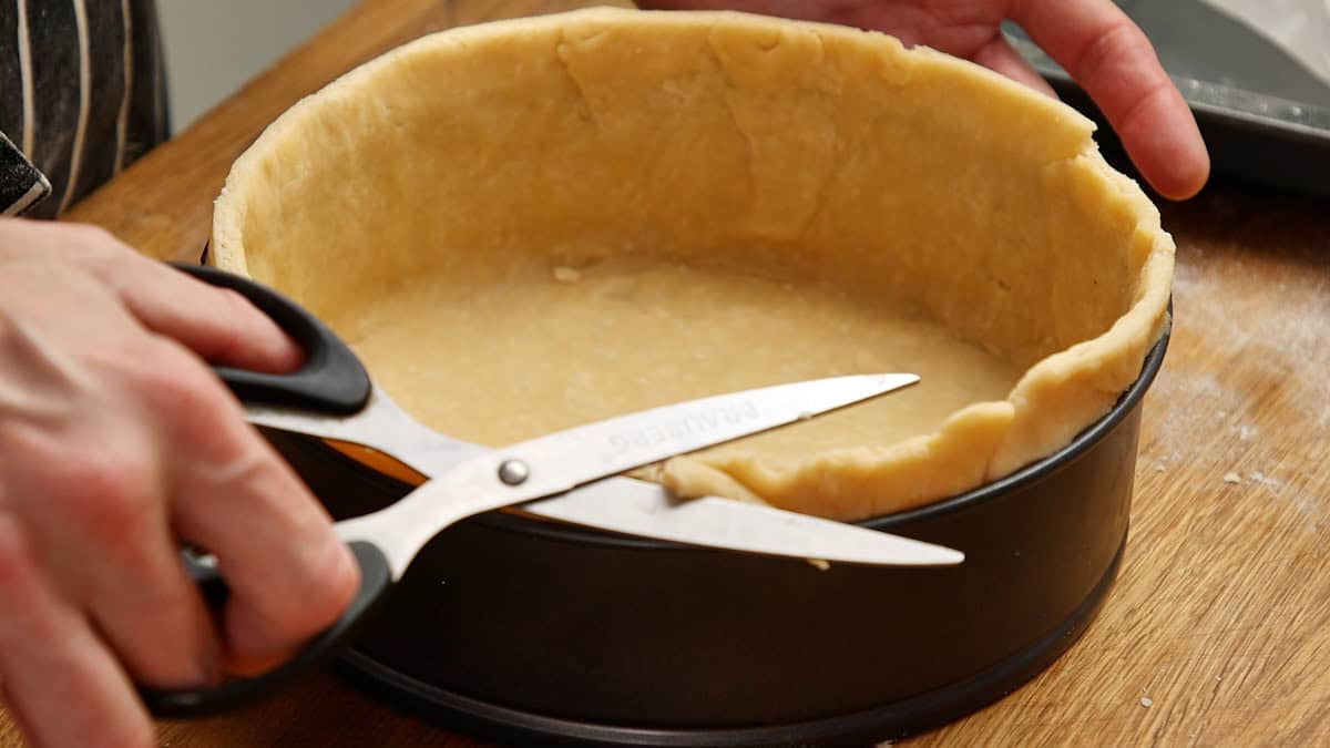 Pastry-lined springform tin being trimmed with kitchen shears.
