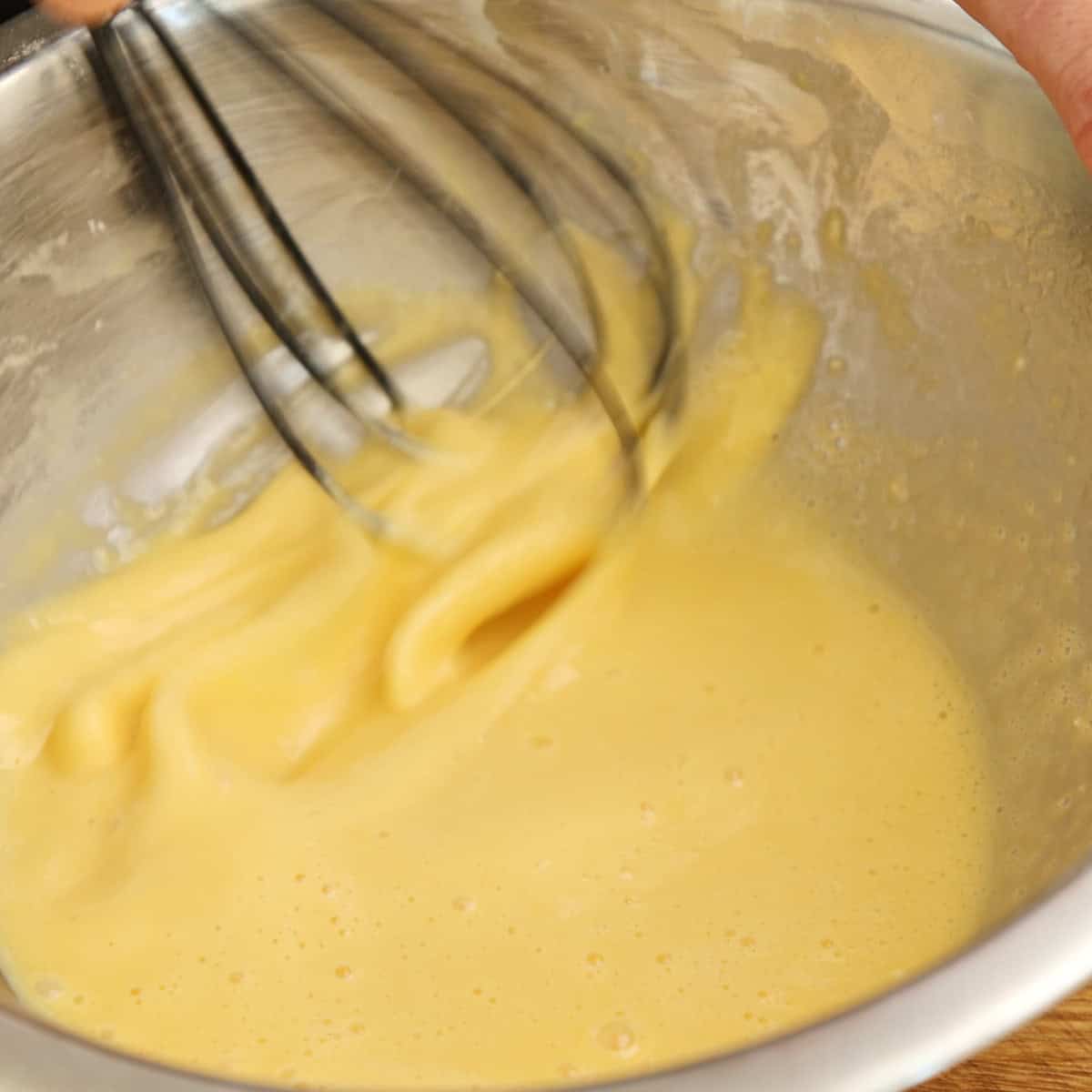 Whisk beating pale yellow custard mixture in a metal bowl.